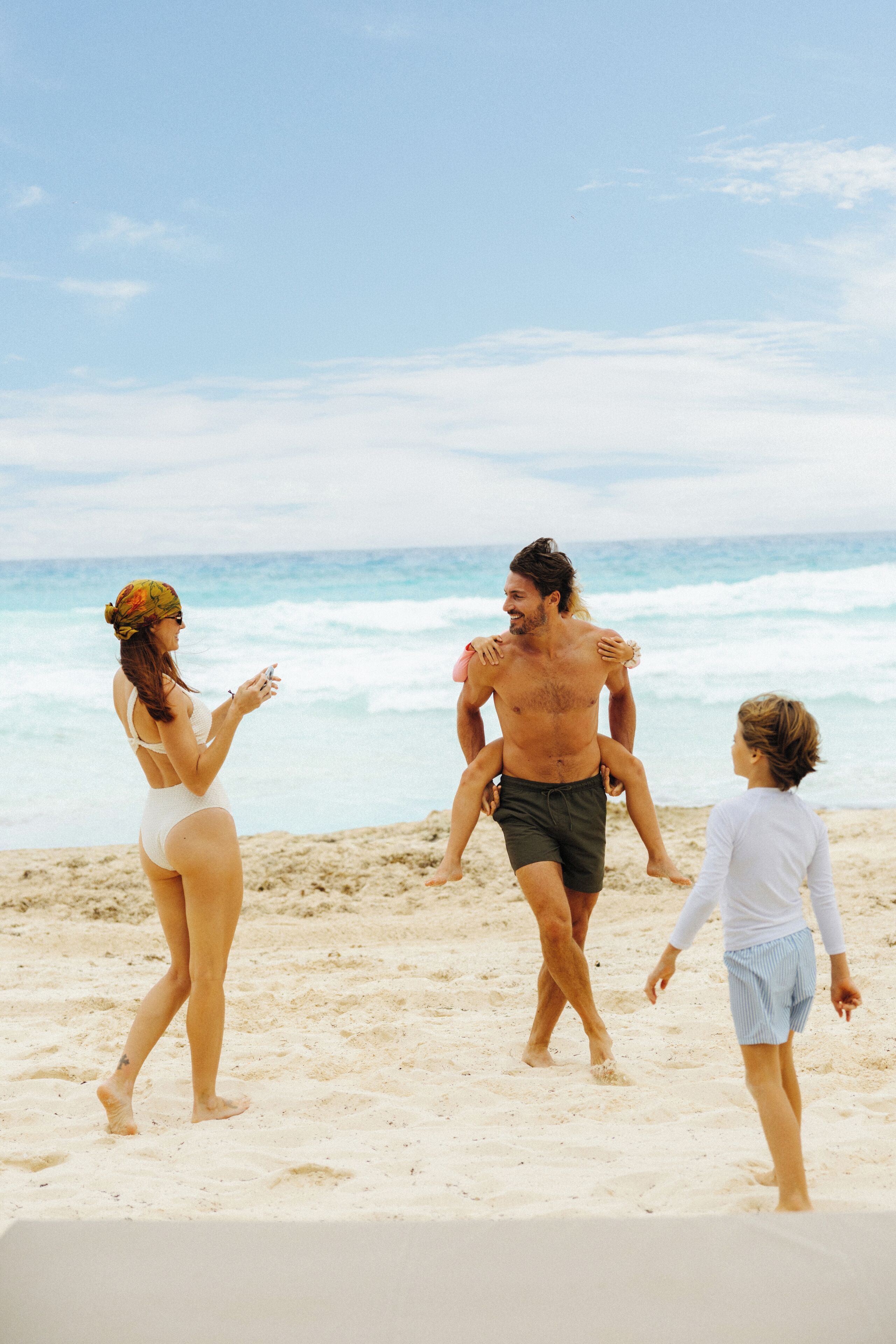 A beach scene with a person carrying another on their shoulders, a child walking beside them, and a woman standing nearby.
