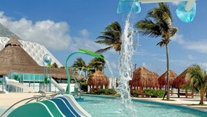 A water slide at a resort with clear blue water, palm trees, and thatched-roof huts. - Paradisus Cancún – All Inclusive (Cancun)