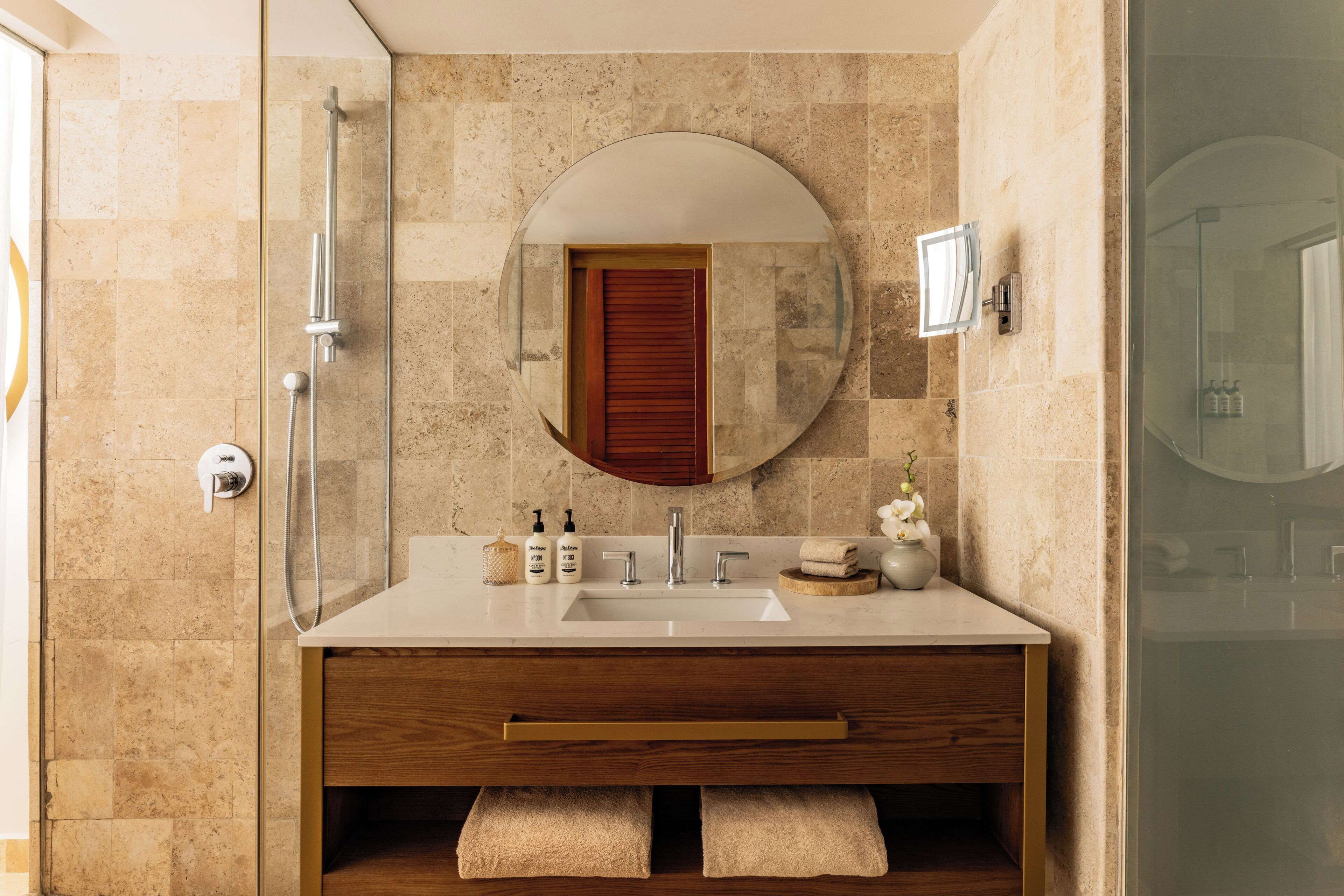 A bathroom with a glass shower enclosure, a round mirror, a wooden vanity with a sink, and a towel rack.
