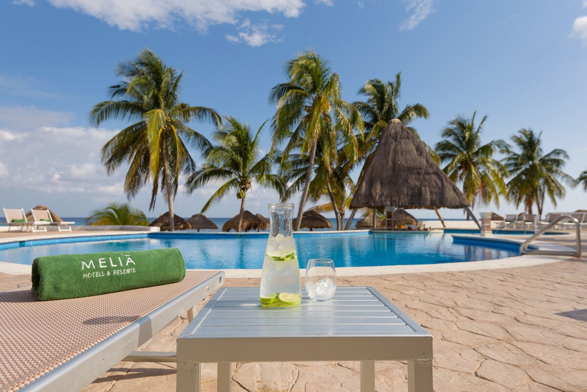 A carafe of a cold citrus beverage sits on a side table next to the pool at Melia Cozumel All Inclusive with a line of palm trees in the background.