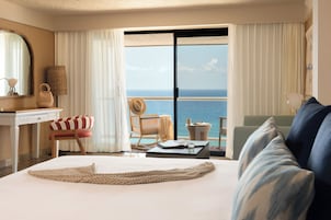 Habitación de hotel con una cama grande, vista al océano, una mesa de comedor con un libro y vasos, y una silla con un sombrero.