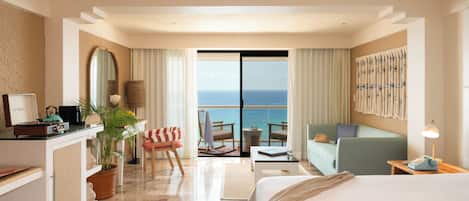 A hotel room with a bed, a desk with a computer, a chair, a small table, a potted plant, and a view of the sea through a glass door.