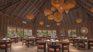 A spacious indoor dining area with multiple tables and chairs, large windows offering views of greenery, and a high ceiling with woven light fixtures. - Zel Cozumel (Cozumel)