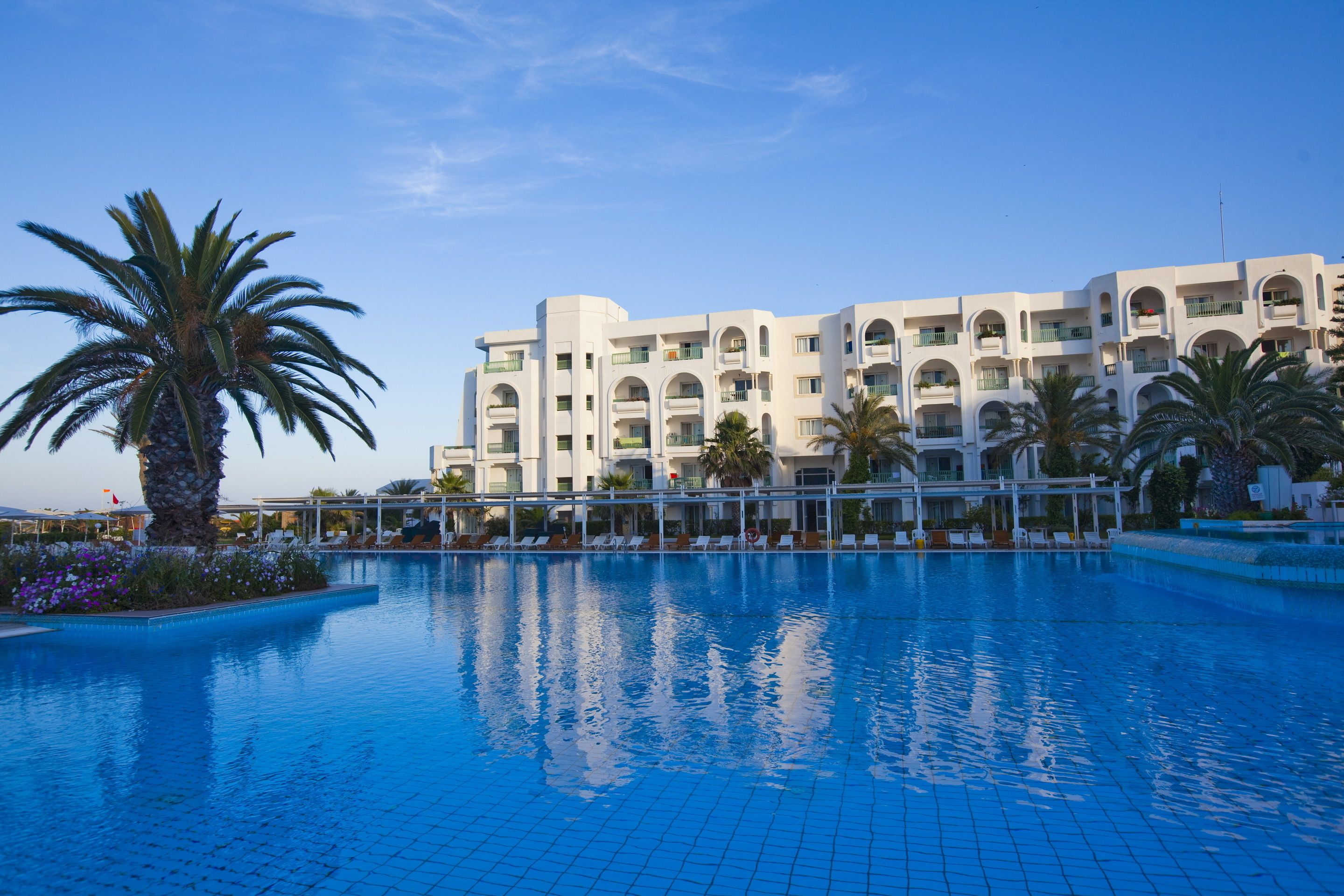 A hotel with a swimming pool, palm trees, and a clear blue sky.
