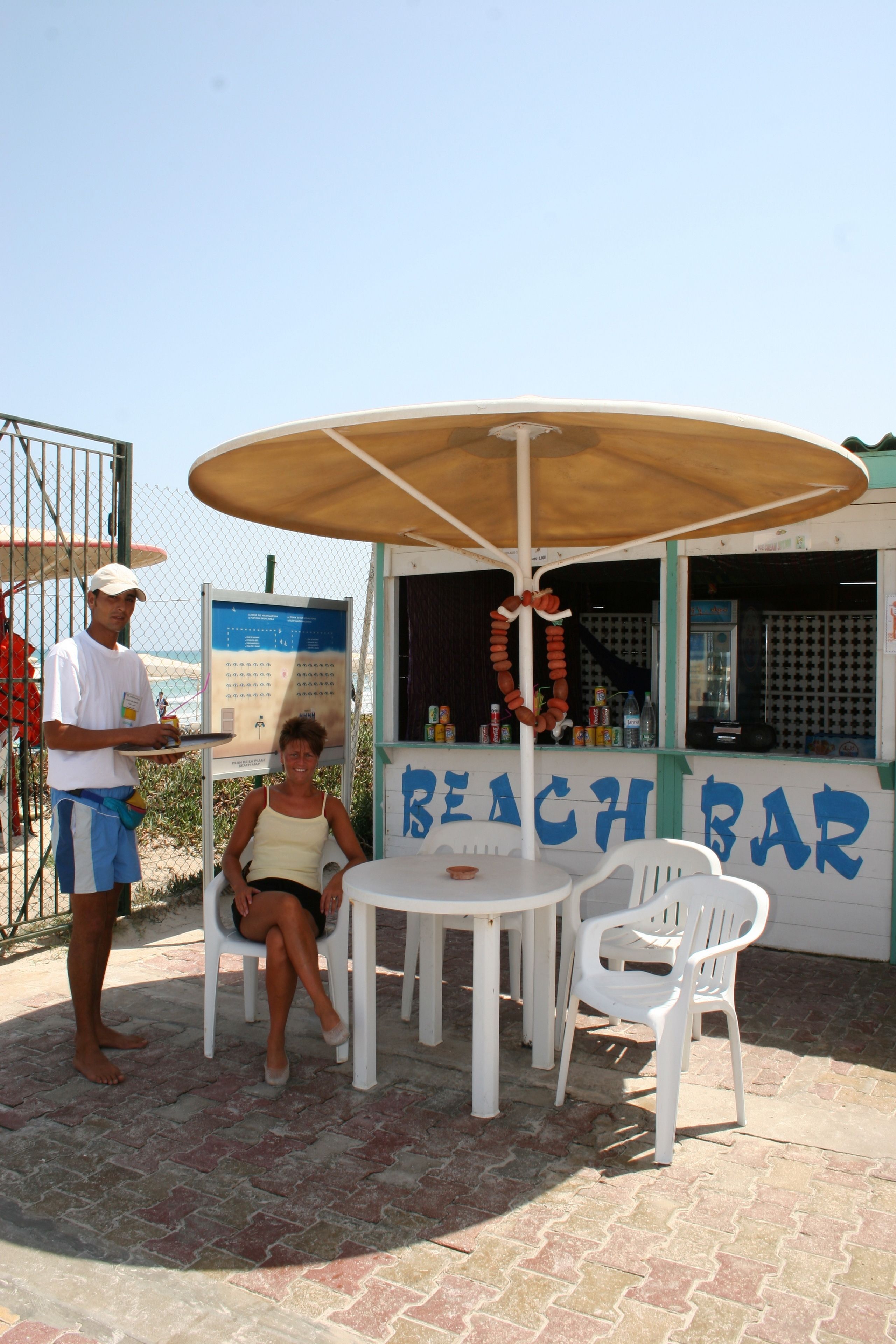 A beach bar with a table and chairs, a menu board, and a person serving food.