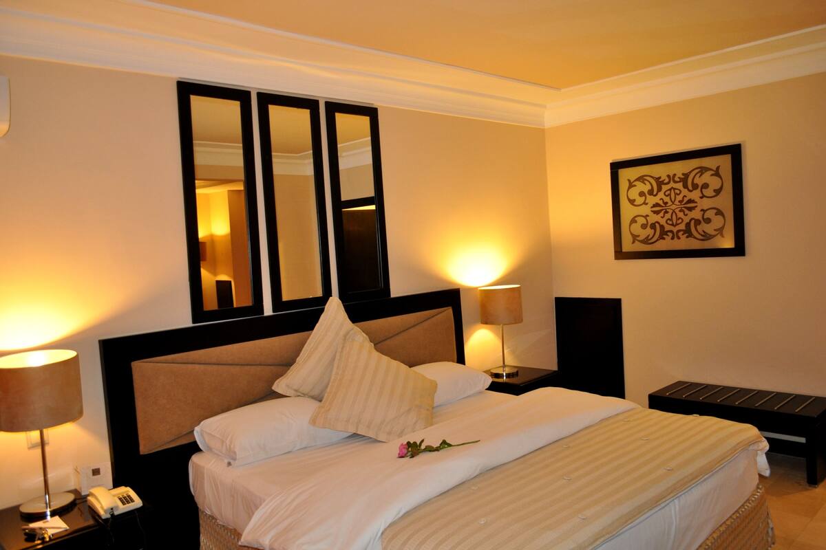 A hotel room with a large bed, two bedside lamps, a bedside phone, a small table, and a decorative wall piece.