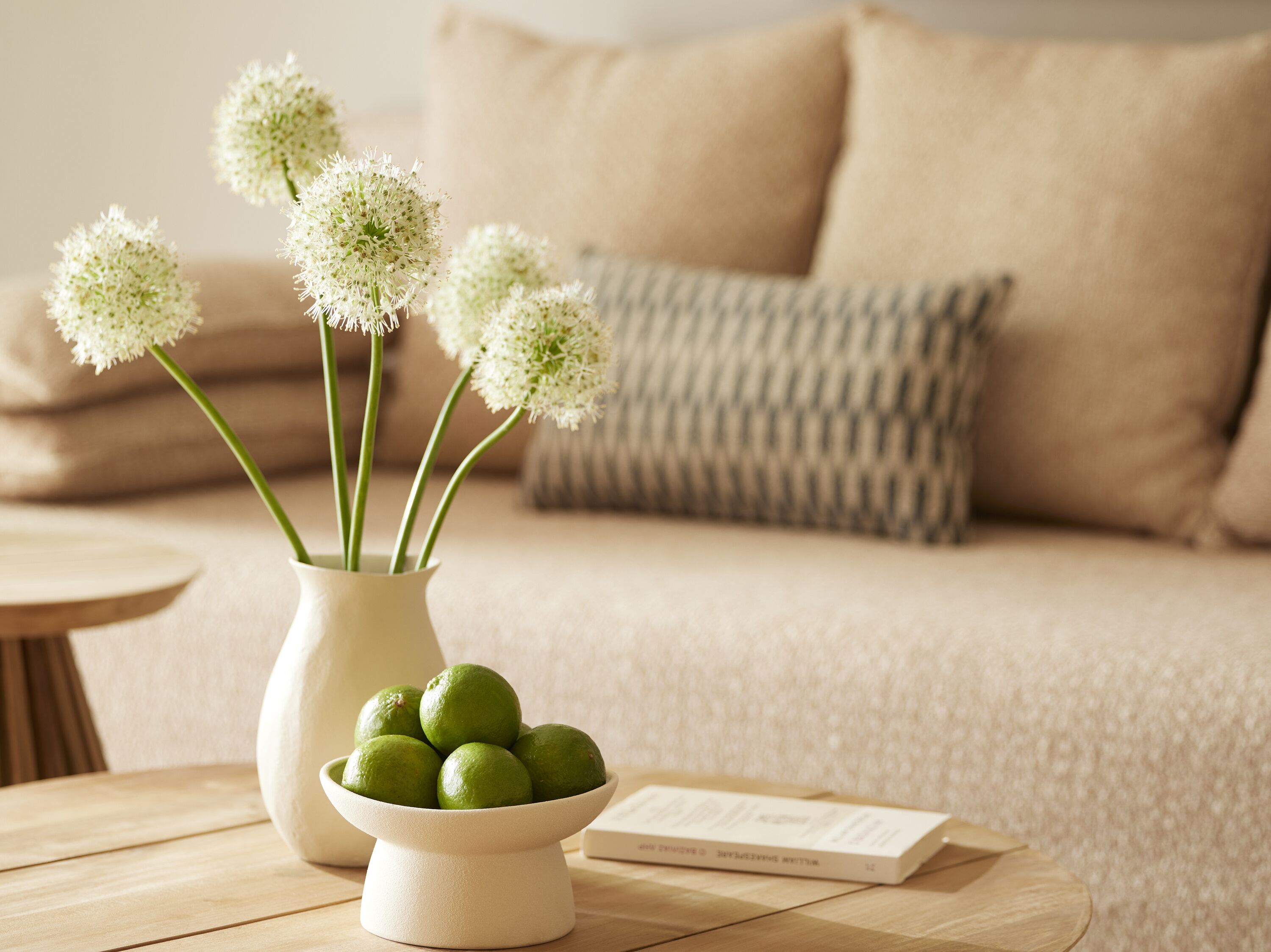 Ein Wohnzimmer mit einem beige farbenen Sofa, einem runden Holztisch, einer weißen Vase mit weißen Blumen und einer Schüssel mit grünen Limetten.