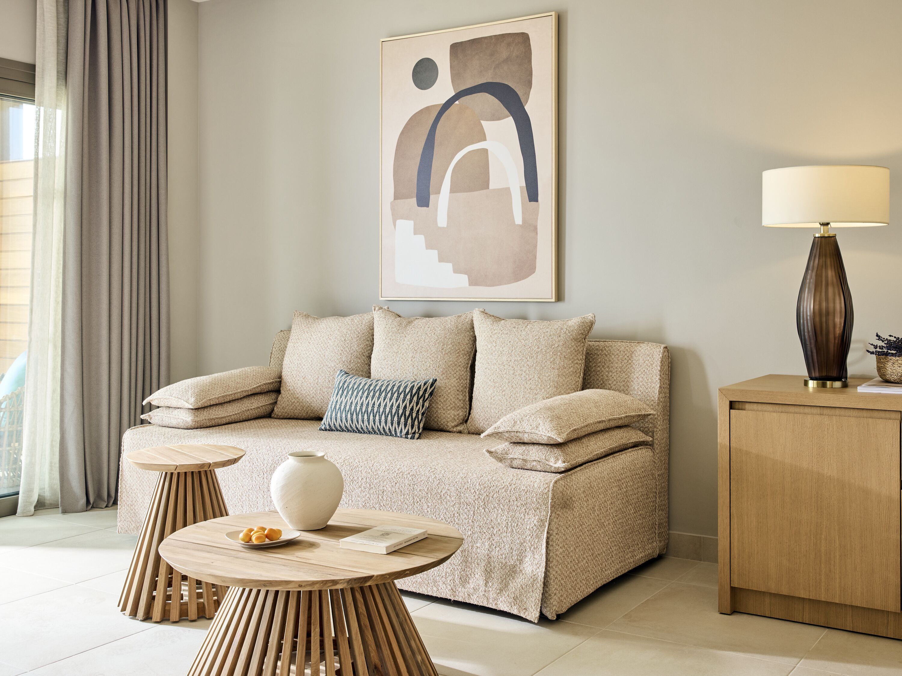 A modern living room with a beige sofa, a round wooden coffee table, a white vase, and a lamp on a wooden side table.