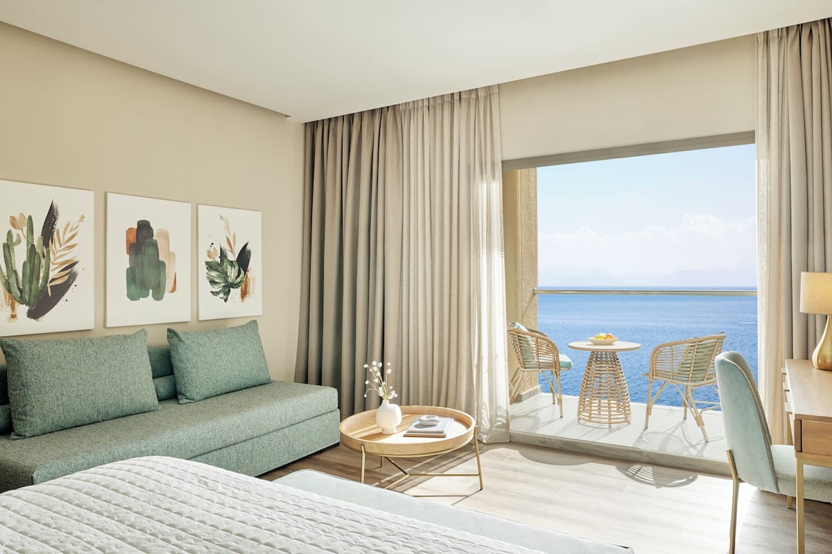 Ein Hotelzimmer mit Bett, einer Couch, einem runden Tisch mit Blumenvase sowie einem Balkon mit Stühlen und Tisch.