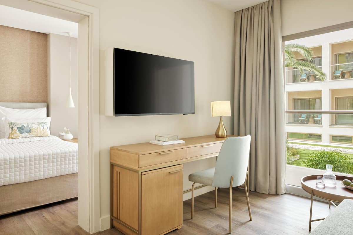 Ein Hotelzimmer mit einem Bett, einem Schreibtisch mit Stuhl, einem Fernseher, einer Lampe und einem Fenster mit Blick auf ein Gebäude und Grünflächen.