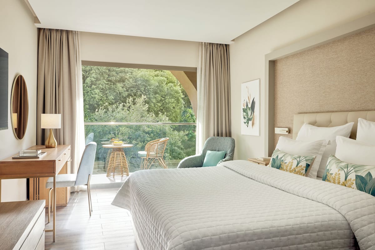 Ein Hotelzimmer mit einem großen Bett, einem Schreibtisch mit Lampe, einem Sessel und einem kleinen Tisch mit Obstschale. Ein Balkon bietet Blick auf Grünflächen.