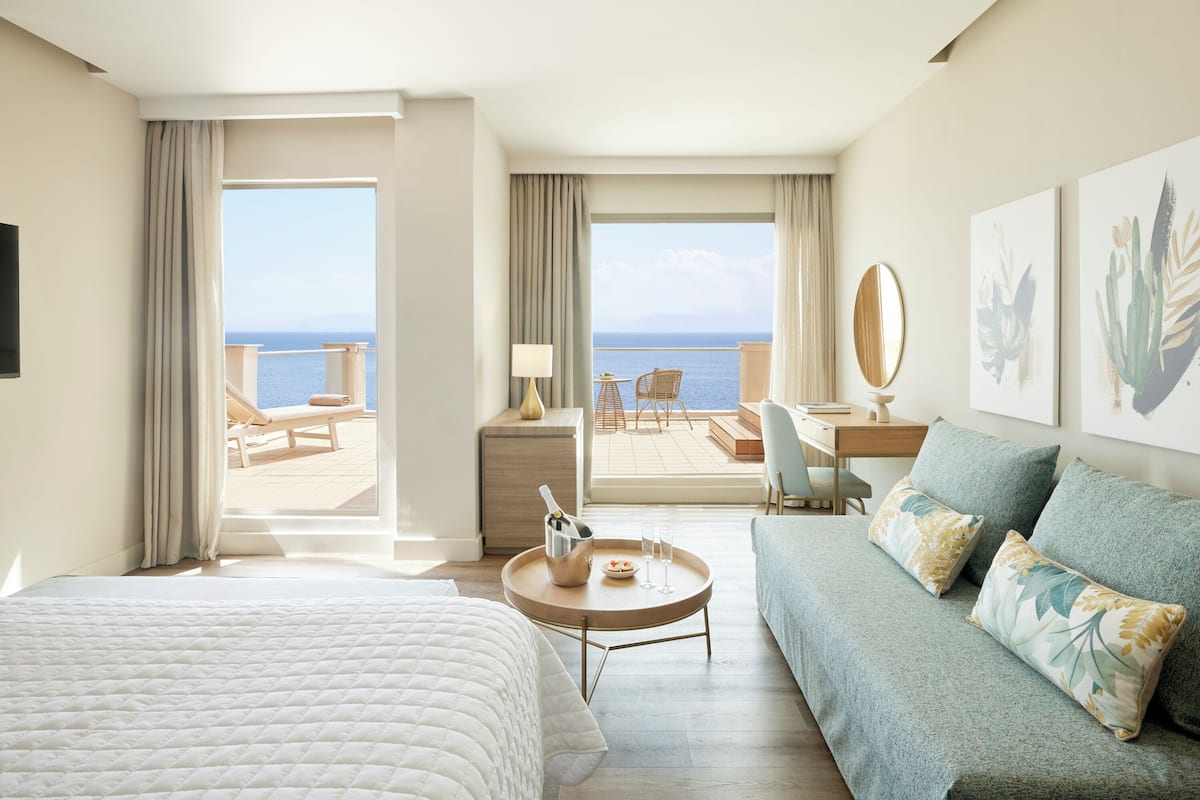Ein Hotelzimmer mit Bett, Sofa, Schreibtisch und einem Balkon mit Meerblick.