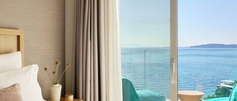 A hotel room with a bed, a view of the sea, and a balcony with chairs.