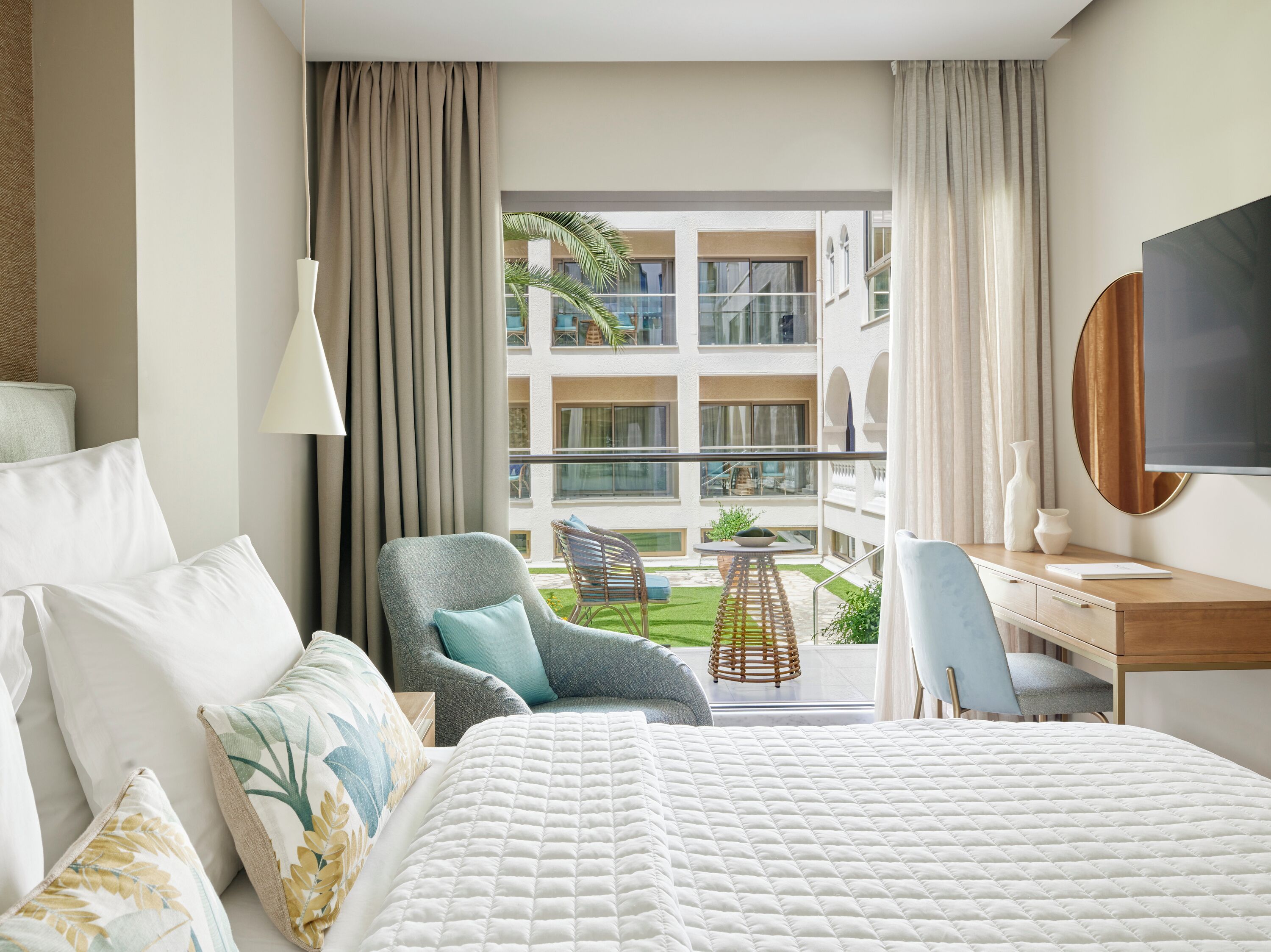 Une chambre d’hôtel comprenant un lit, deux chaises, un bureau, une télévision et un balcon avec vue sur les bâtiments et la verdure.