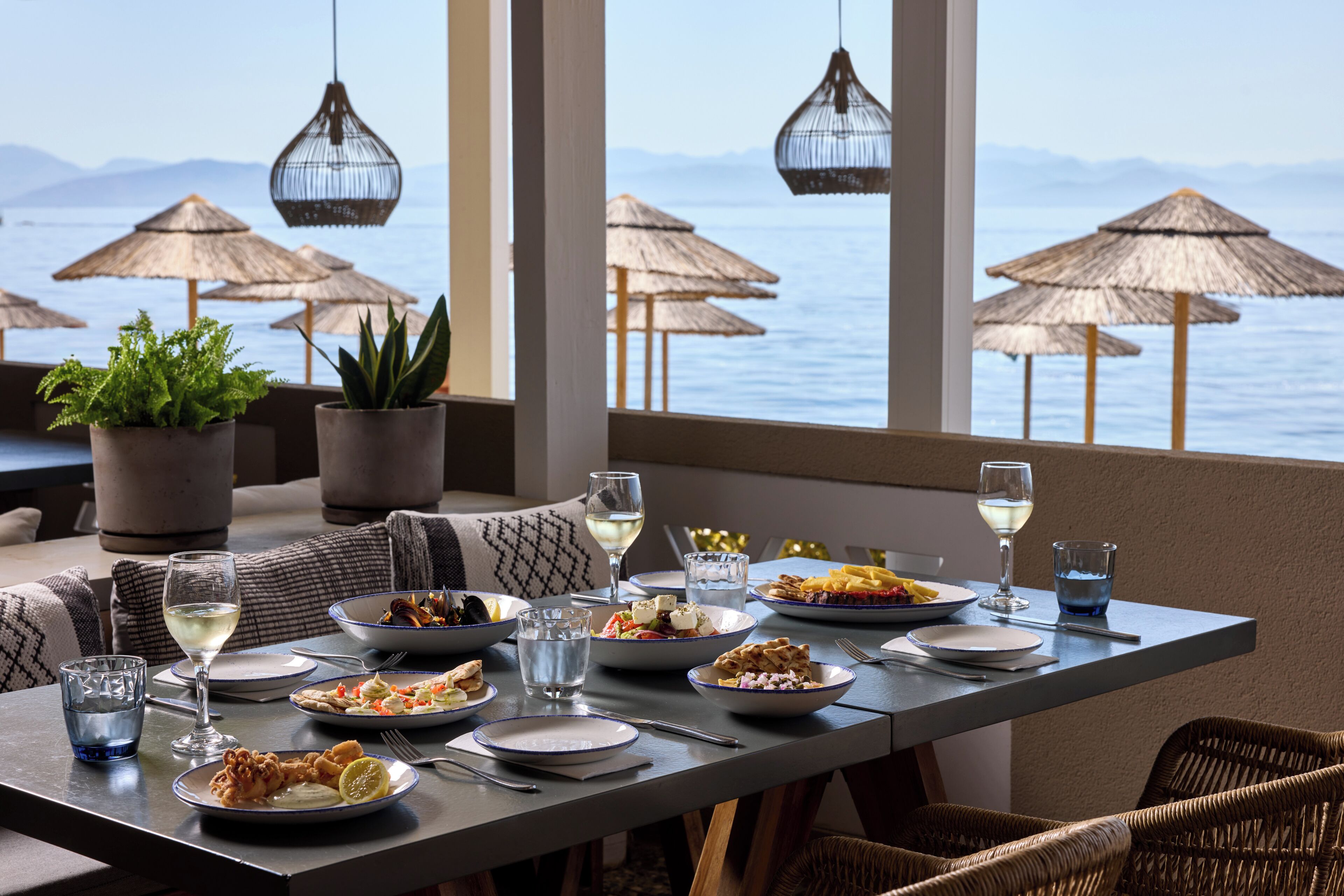 A dining table set with various dishes, glasses, and cutlery, overlooking a body of water with thatched umbrellas.