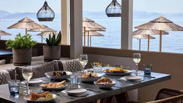A dining table set with various dishes, glasses, and cutlery, overlooking a body of water with thatched umbrellas.