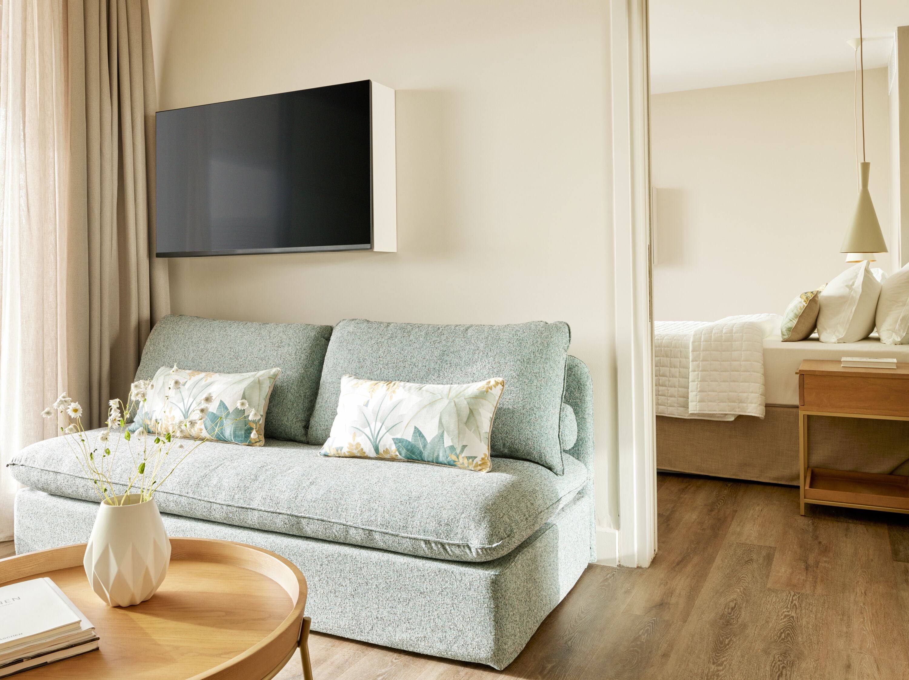 Un salón moderno con un sofá gris, una mesita de madera, un jarrón blanco con flores y un televisor de pantalla plana montado en la pared.