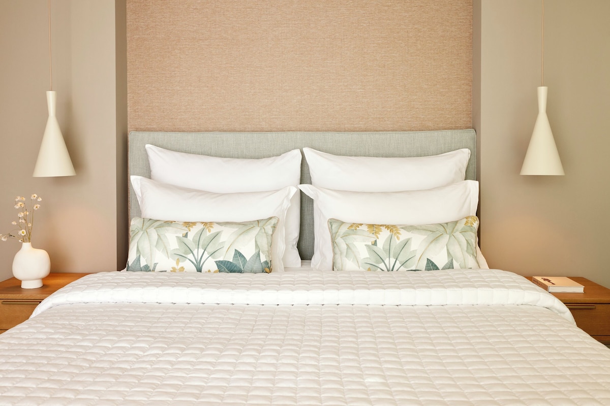 Ein ordentlich bezogenes Bett mit weißer Steppdecke, ein Kopfteil mit einer strukturierten, beige farbenen Zierleiste und zwei Nachttischlampen.