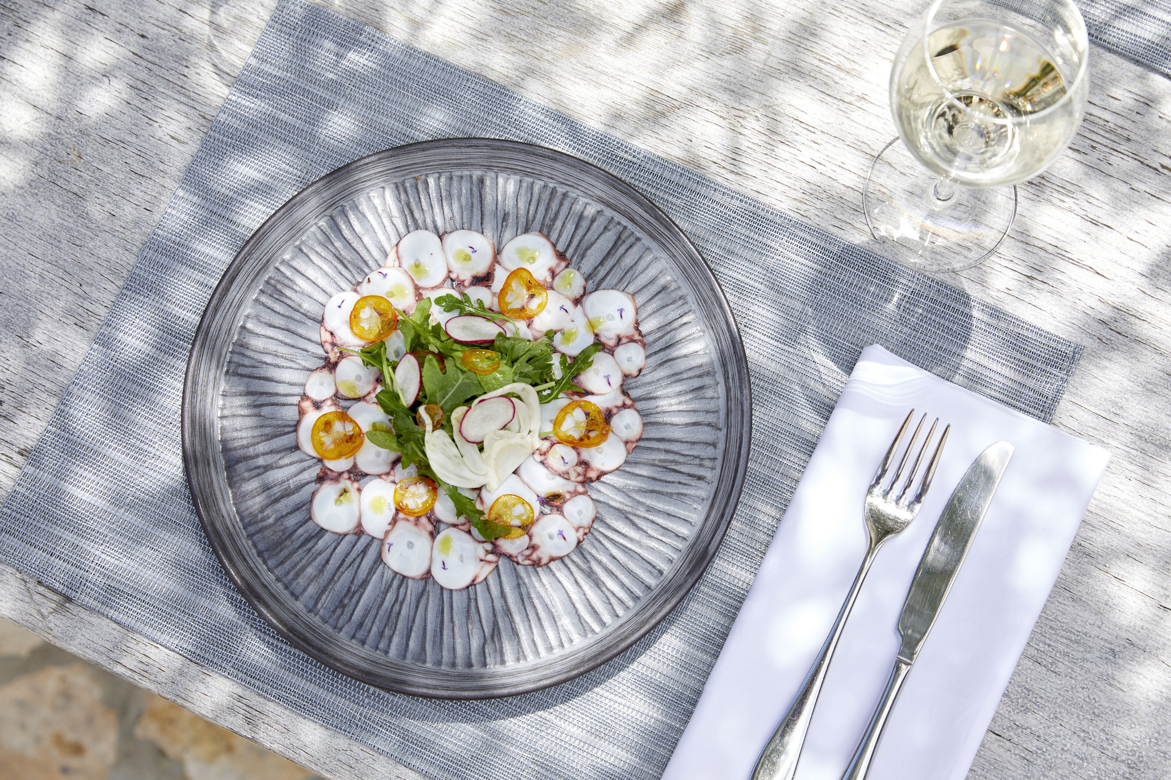 A clear glass plate with a salad on a table, accompanied by a fork and knife on a napkin.