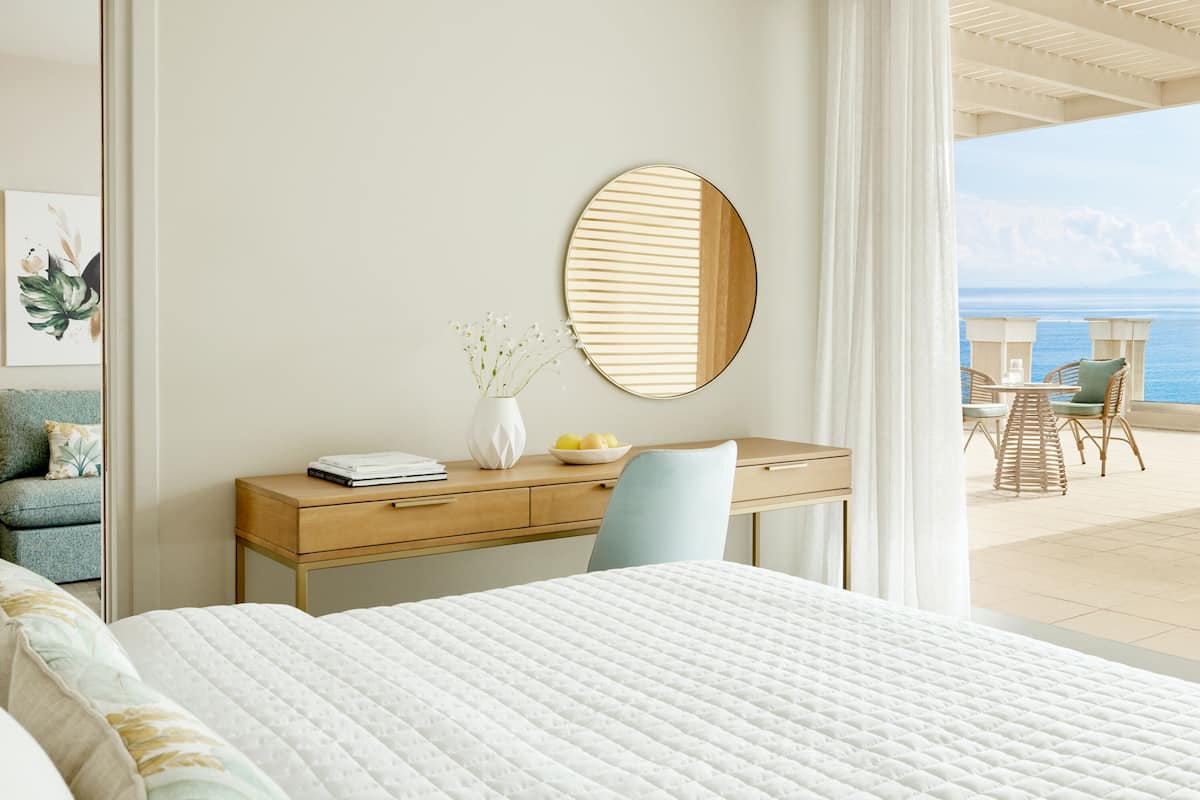 Ein Schlafzimmer mit Bett, Schreibtisch mit Stuhl, rundem Spiegel und Meerblick vom Balkon.