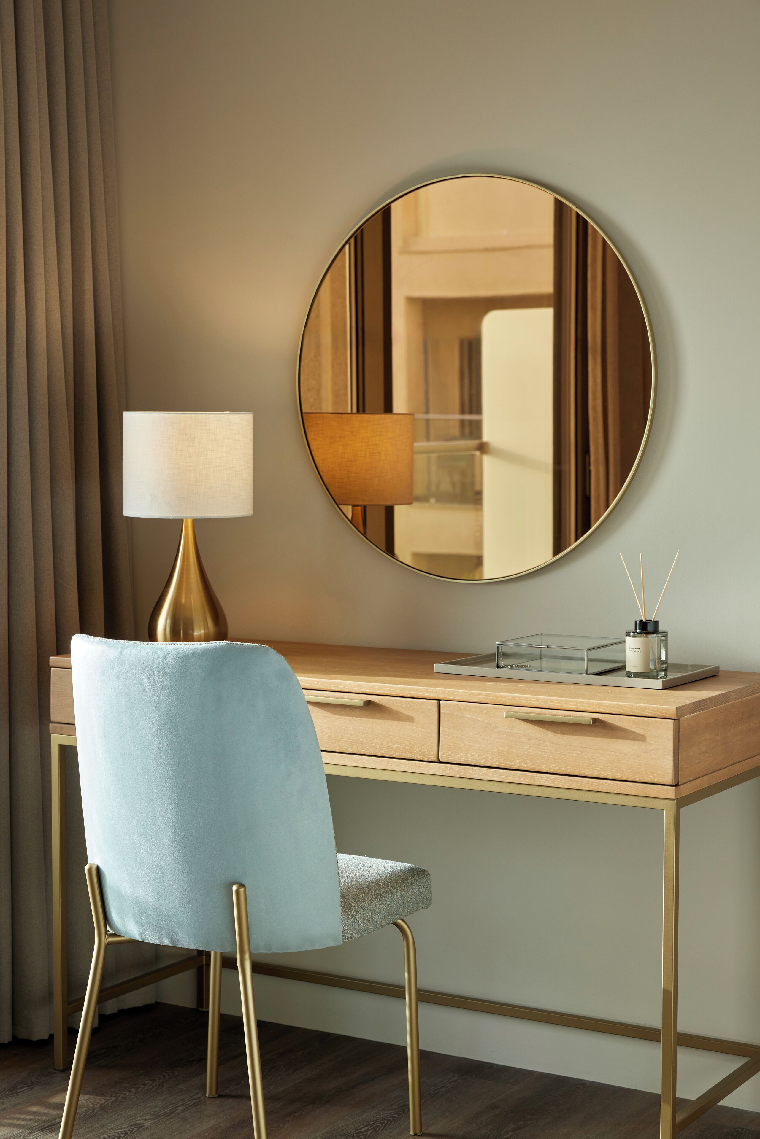 A hotel room with a wooden desk, a round mirror, a lamp, and a chair.