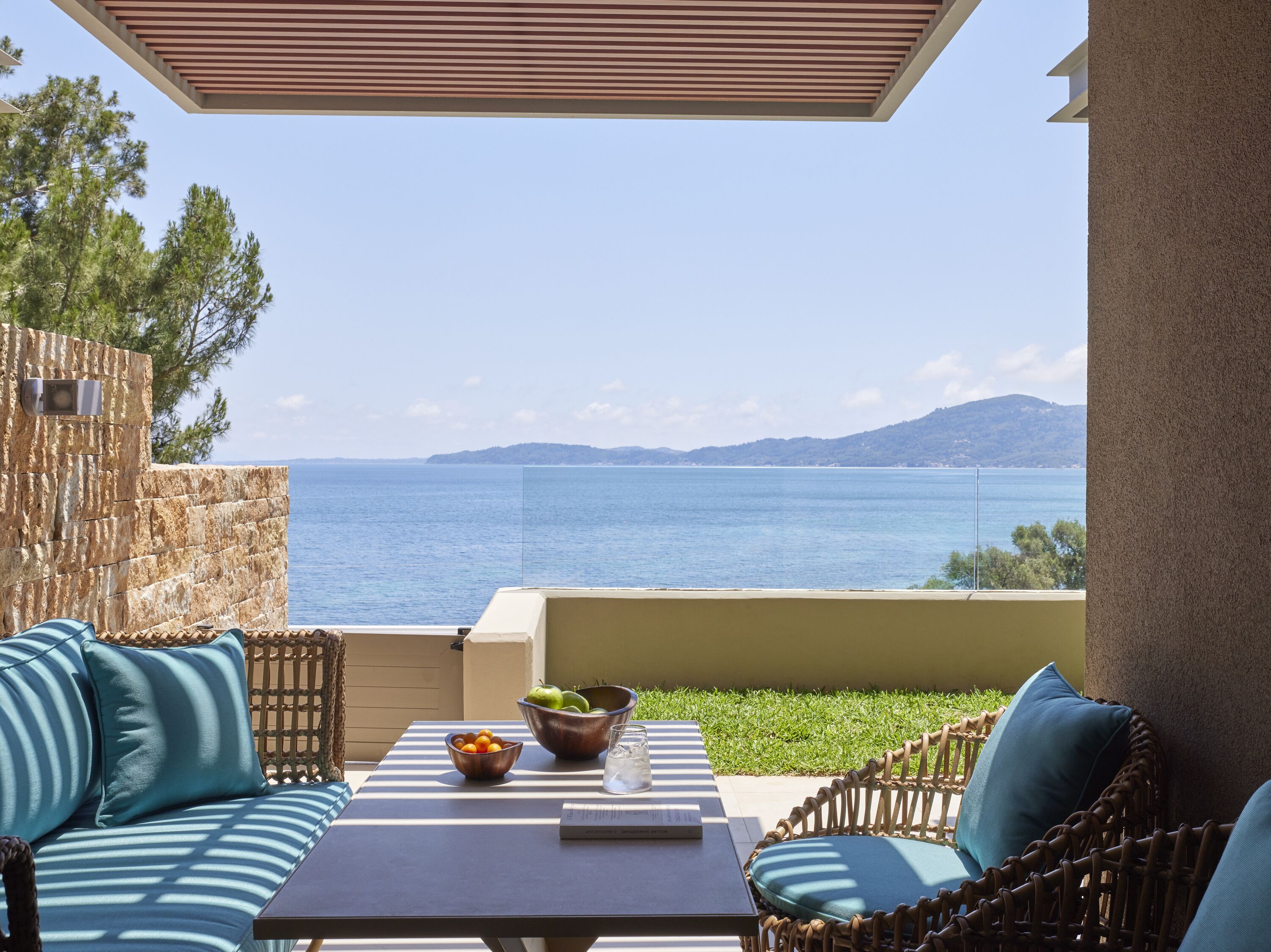A balcony with wicker furniture, a table with fruit and a glass, and a view of the sea and hills.