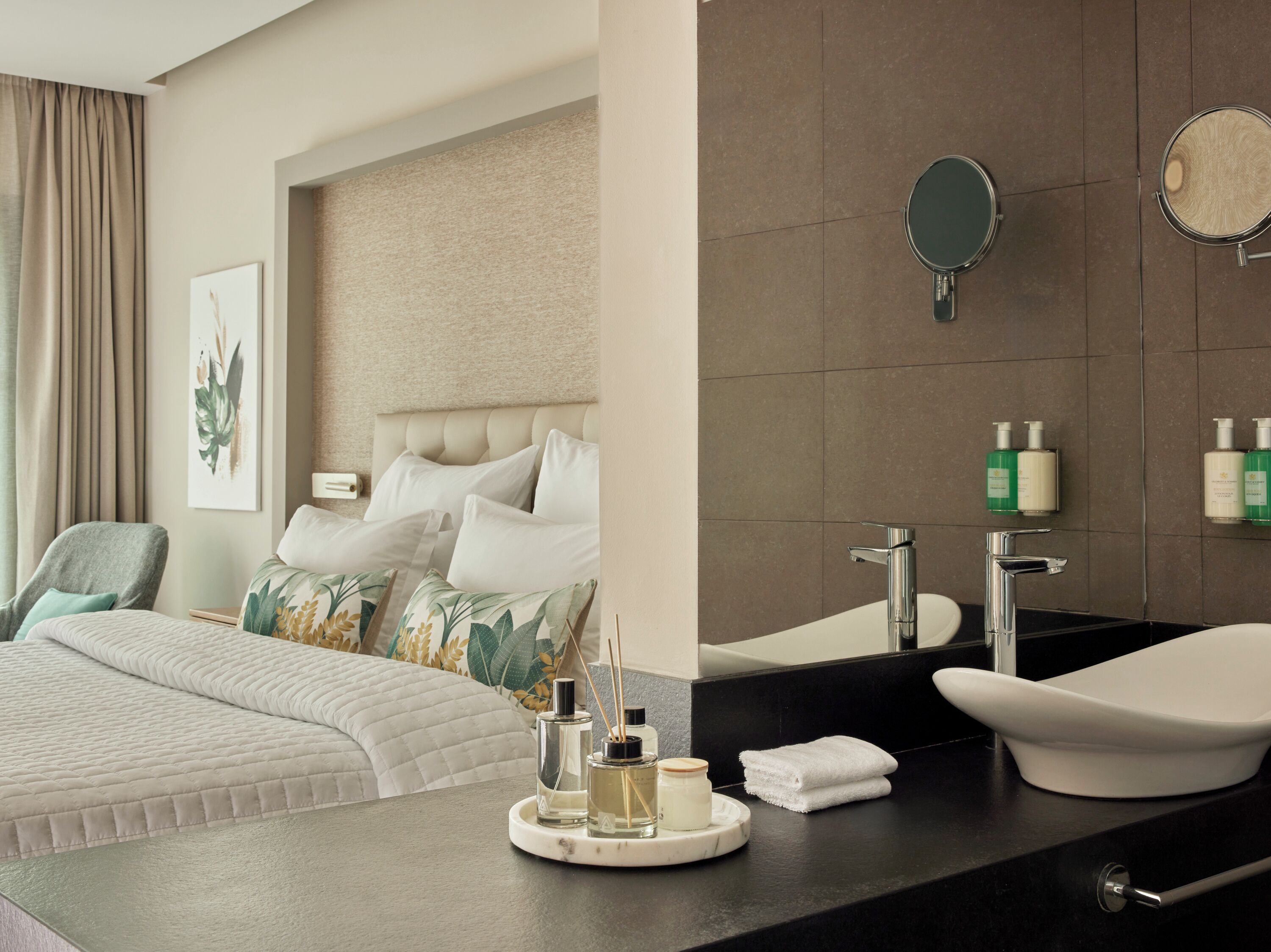 A modern hotel room with a large bed, a bedside table with toiletries, a bathroom with a vanity and mirror, and a large window with curtains.