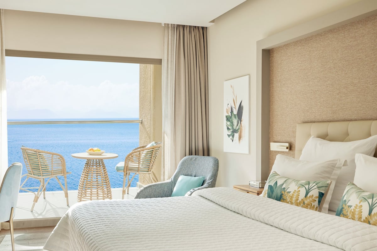 Ein Hotelzimmer mit einem großen Bett, Meerblick, einem Balkon mit Korbstühlen und einem Bild an der Wand.