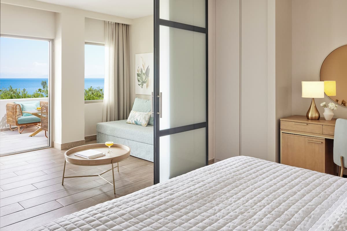 Ein Hotelzimmer mit Bett, rundem Tisch mit Getränk, Sofa mit Aussicht und Schreibtisch mit Lampe.