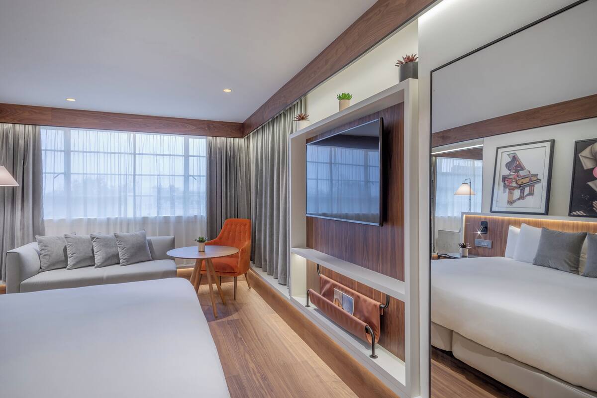 A hotel room with a large bed, a sofa, a small table, a TV mounted on the wall, and a window with curtains.