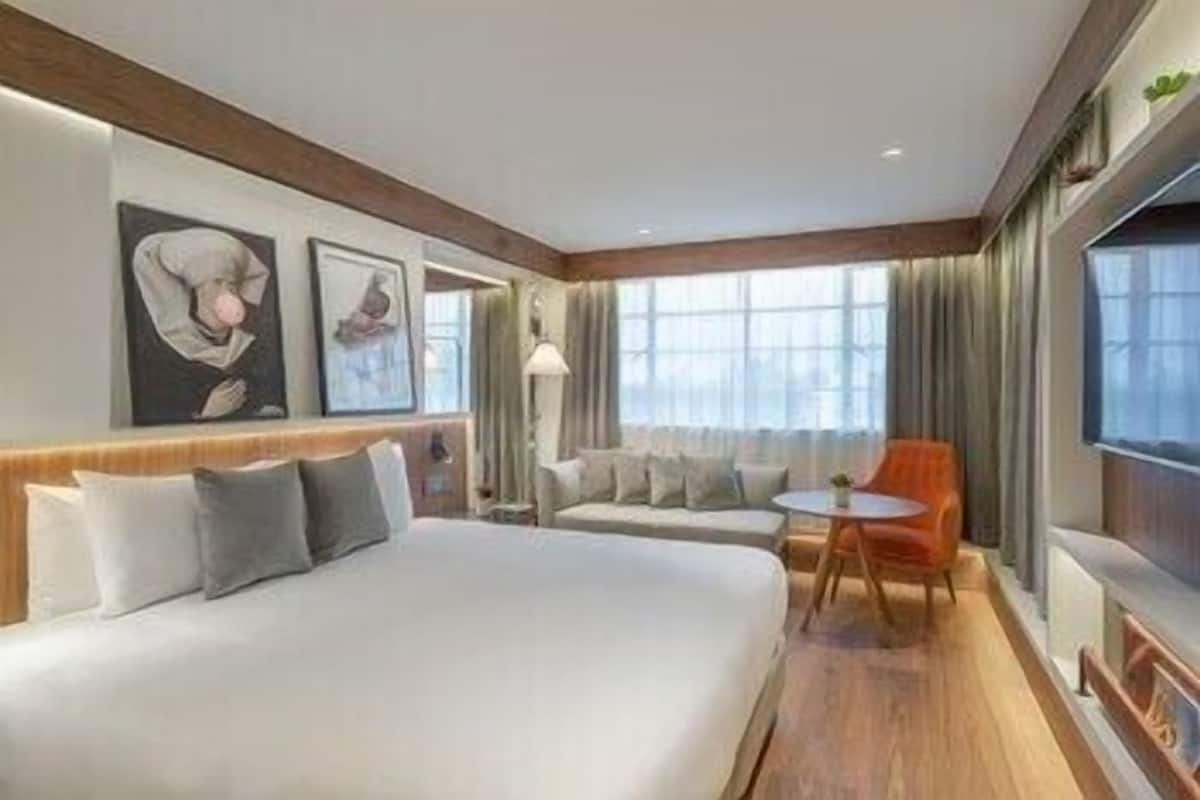 A hotel room with a large bed, a sofa, a small table, and a chair. There are two windows with curtains, a wall-mounted TV, and artwork on the wall.