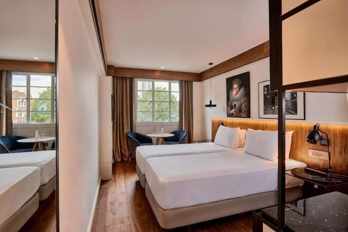 A hotel room with a large bed, a smaller bed, a desk, a chair, and a window with curtains.