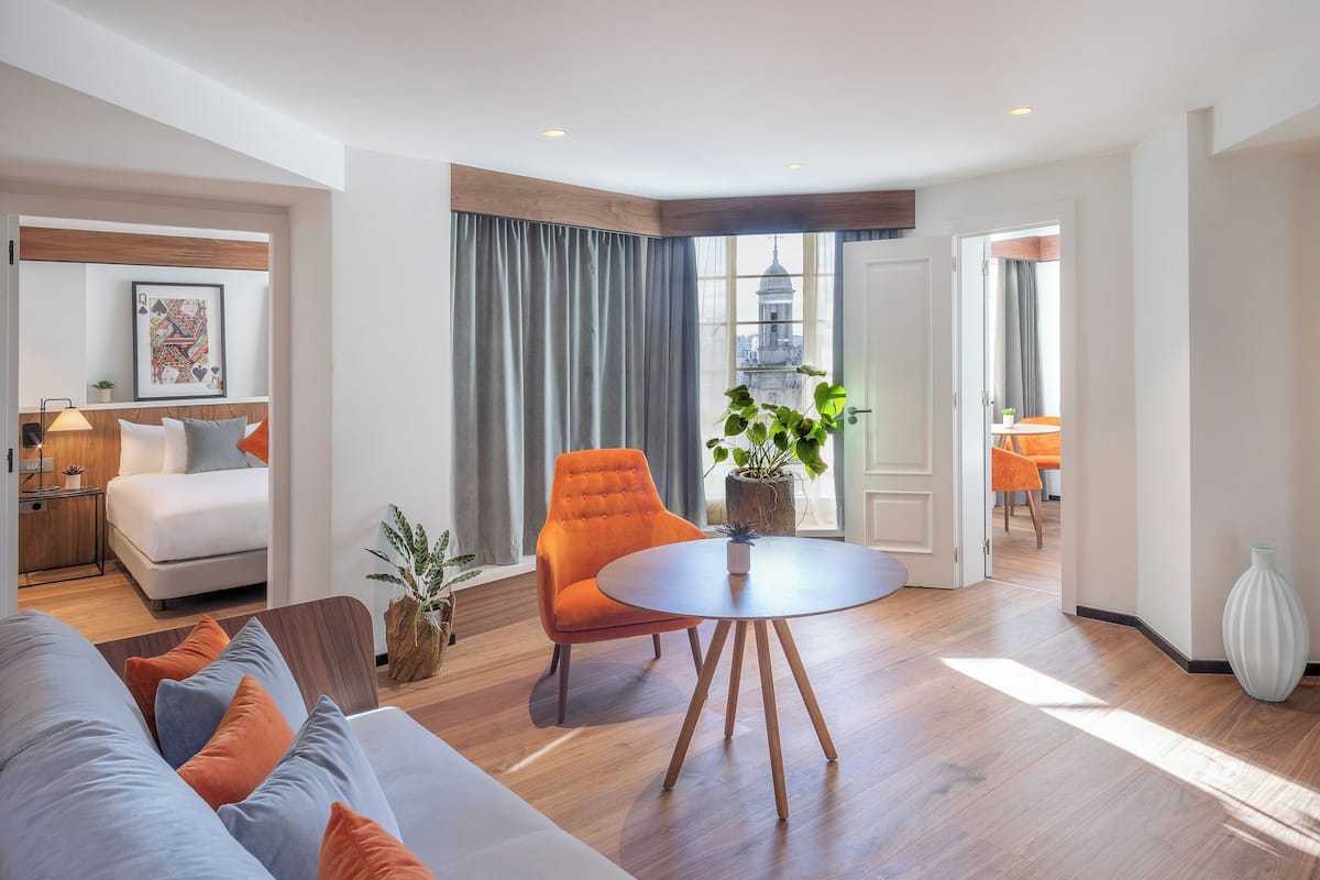 A modern living room with a sofa, a round table, and an orange chair. There is a window with curtains, a plant, and a view of a building.