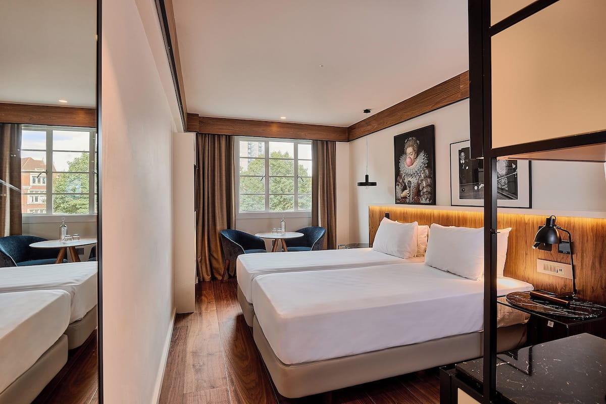A hotel room with a large bed, a desk, two chairs, a small table, and a window with curtains.