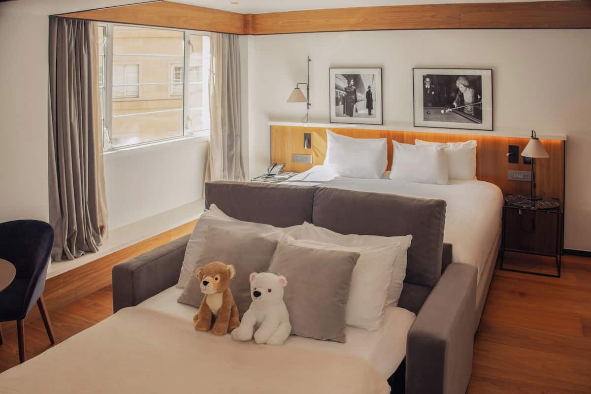 A hotel room with a bed, a sofa, a chair, two teddy bears, a lamp, and two framed pictures on the wall.