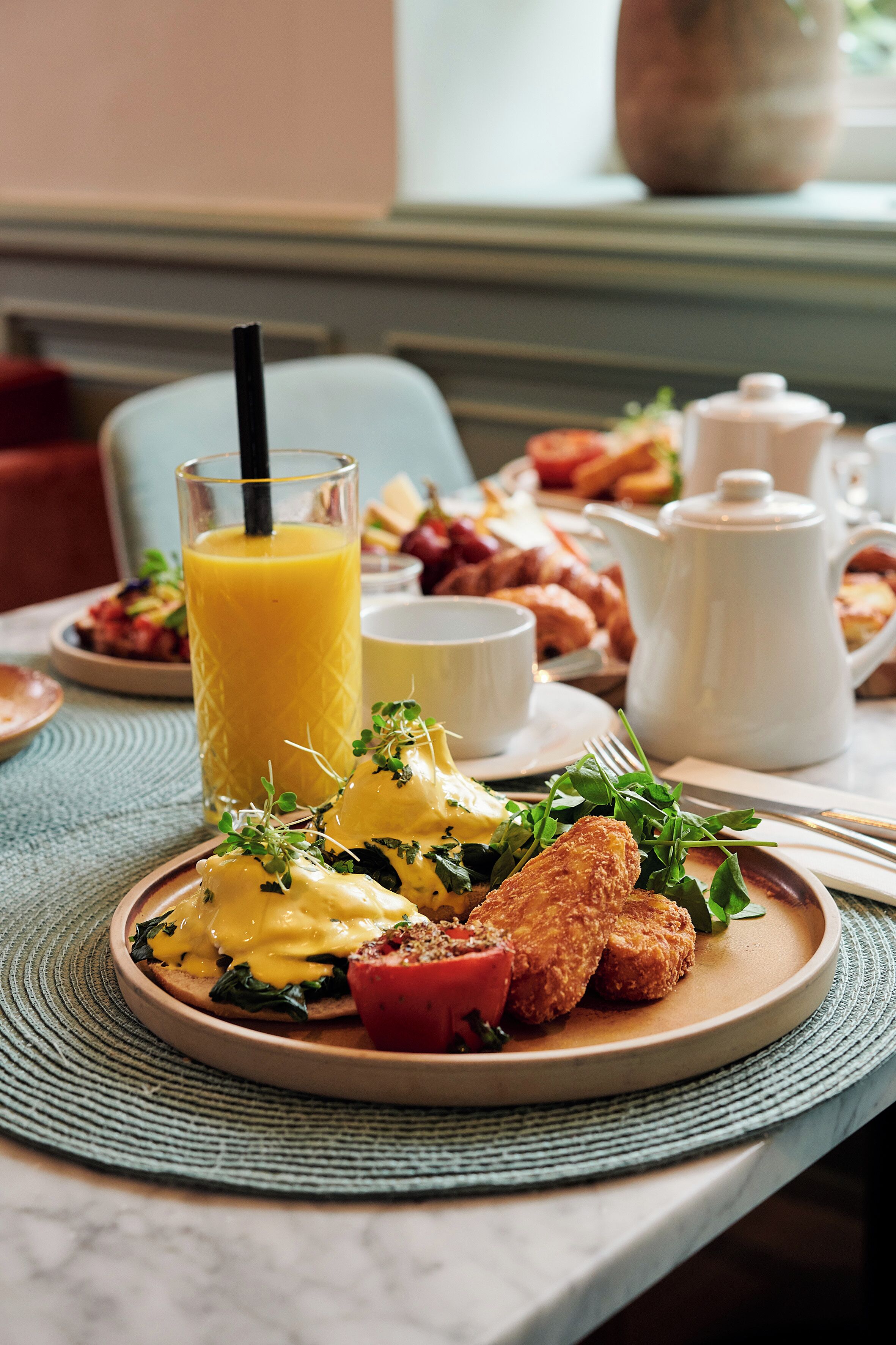 A breakfast setting with a plate of food, a glass of orange juice, a teapot, and a cup.