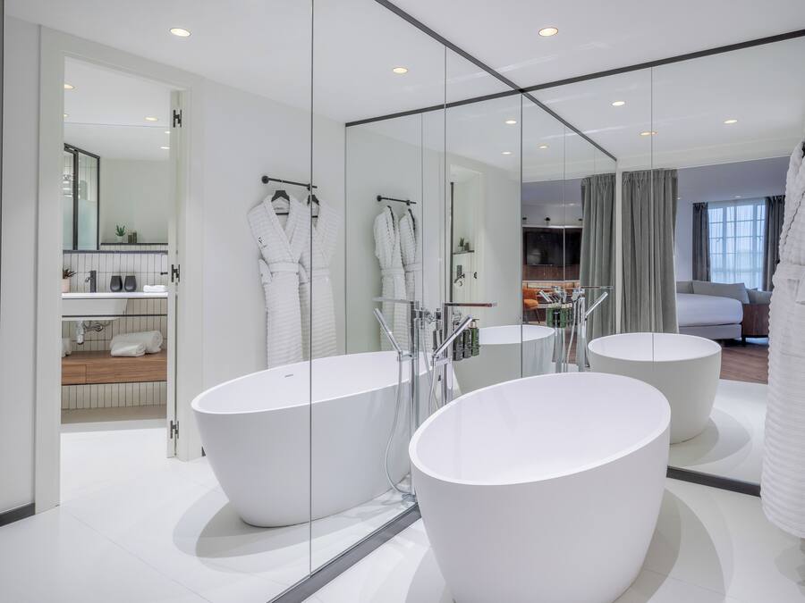 Un baño moderno con dos tinas blancas grandes, una zona de ducha con paredes de vidrio y una cocina visible al fondo.