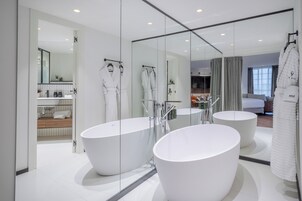 Un baño moderno con dos tinas blancas grandes, una zona de ducha con paredes de vidrio y una cocina visible al fondo.