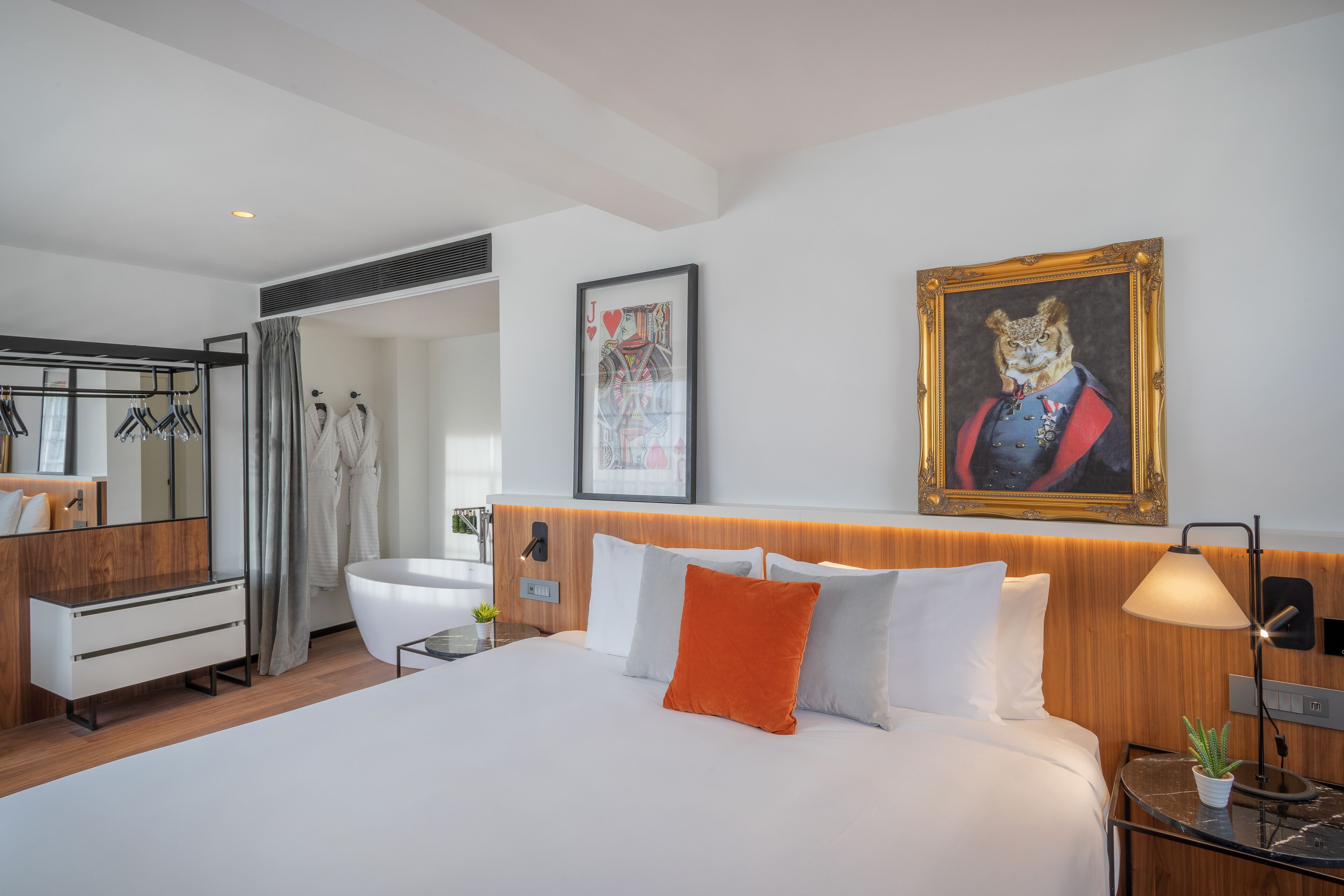 Habitación de hotel moderna con una cama grande, cabecera de madera, un cuadro de un gato con uniforme militar, una foto enmarcada de una carta de juego y un baño con ducha y espejo.