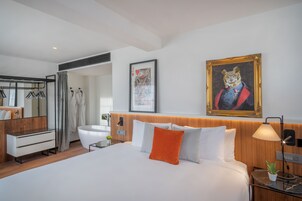 Ein modernes Hotelzimmer mit einem großen Bett, einem Holzkopfende, einem Gemälde einer Katze in Militäruniform, einem gerahmten Bild einer Spielkarte sowie einem Badezimmer mit Dusche und Spiegel.