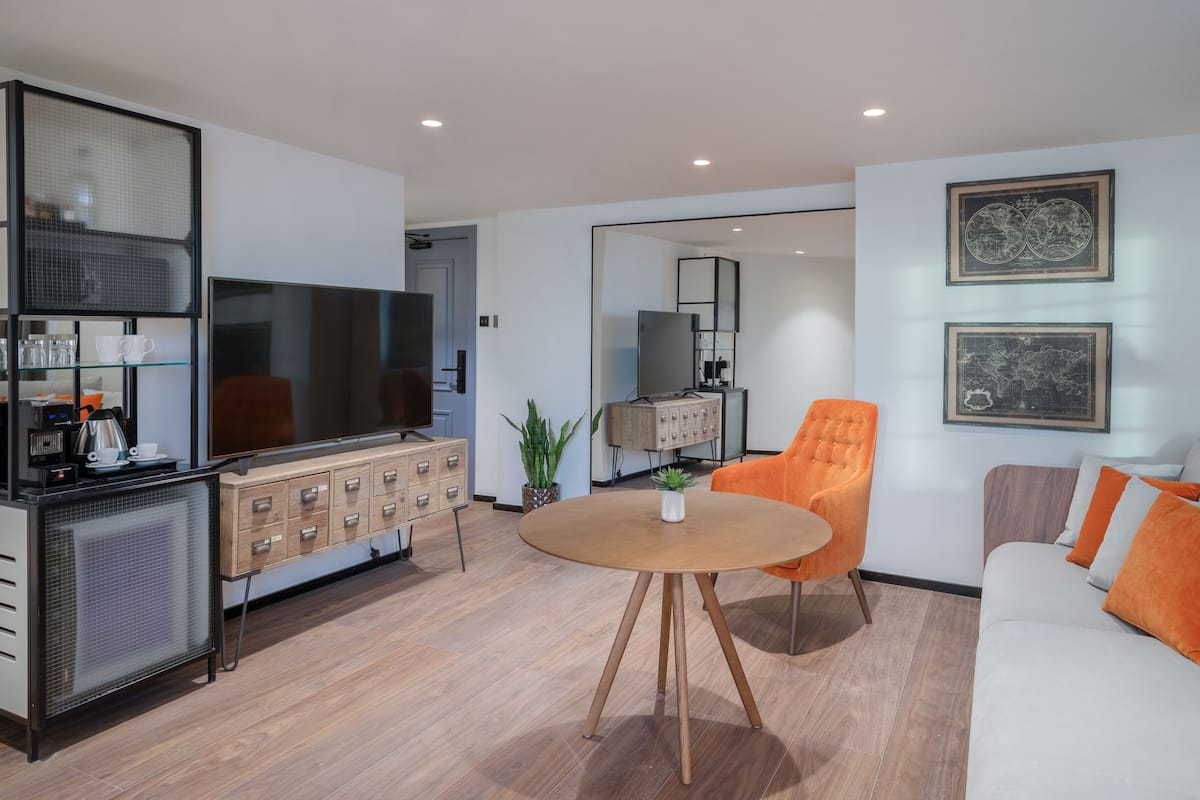 A modern living room with a flat-screen TV, a wooden cabinet, a dining table, and a sofa with orange cushions.
