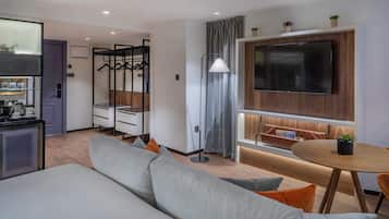 A modern hotel room with a sofa, a dining table, a TV mounted on the wall, and a small kitchen area.