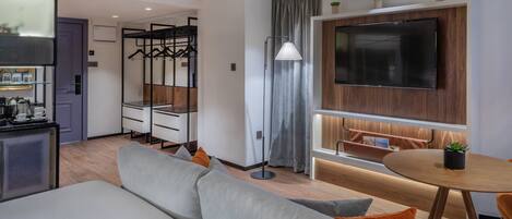 Una habitación de hotel moderna con sofá, una mesa de comedor, un televisor montado en la pared y una pequeña cocina.