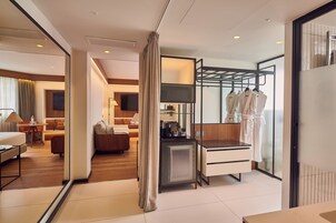 A modern hotel room with a kitchenette, a seating area with a sofa and armchair, a dining table, and a large mirror reflecting the room.