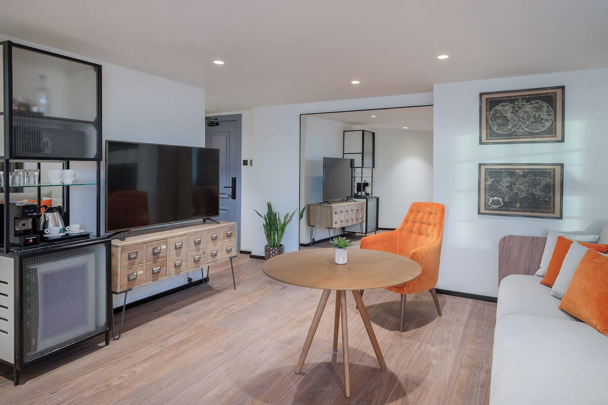 A modern living room with a flat-screen TV, a wooden cabinet, a dining table, and a sofa with orange cushions.
