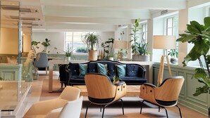 Un amplio salón con un sofá de color oscuro, dos sillas de madera y una mesa de centro. Hay varias plantas en macetas y un ventanal que proporciona luz natural.