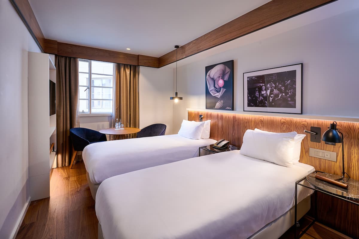 A hotel room with two beds, a desk, a chair, a window with curtains, and wall-mounted artwork.