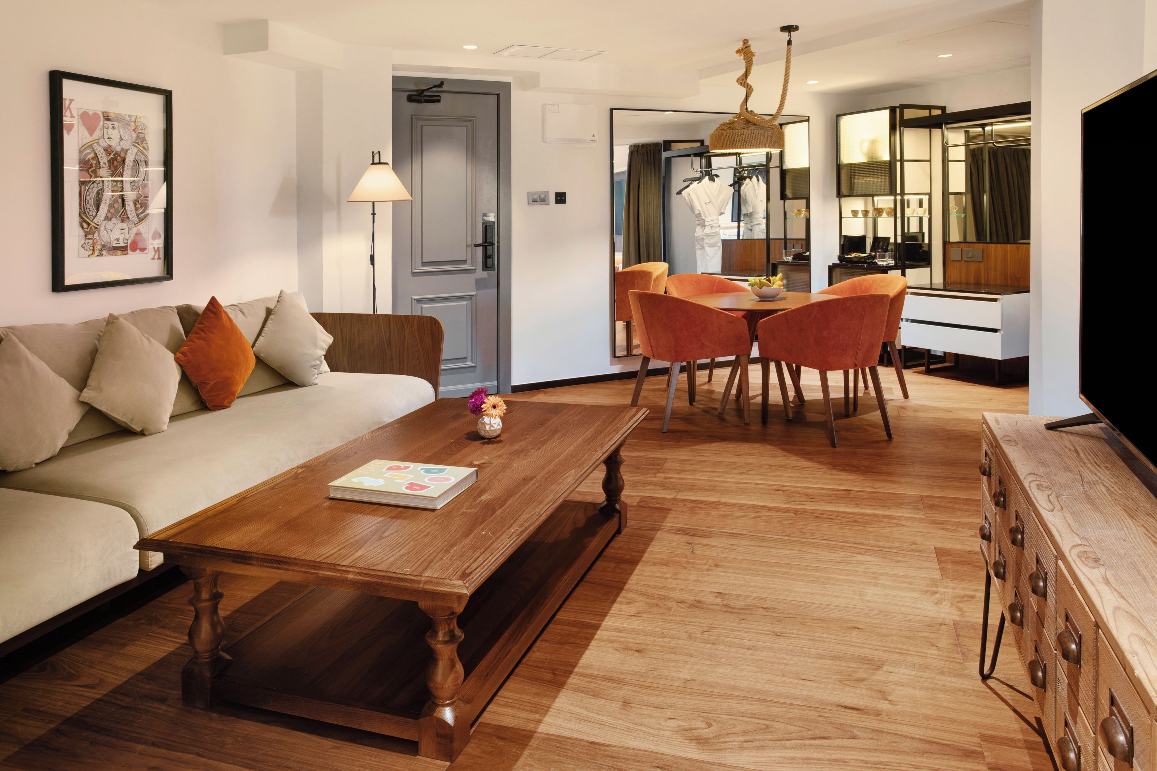 Ein modernes Wohnzimmer mit einer Couch, einem Couchtisch und orangefarbenen Sesseln. Es gibt einen Essbereich mit Tisch und Stühlen sowie eine Küchenzeile mit Kühlschrank und Mikrowelle.
