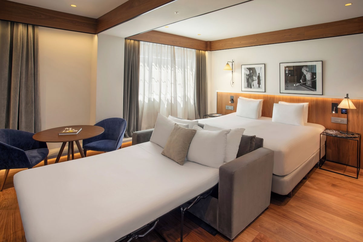 Una habitación de hotel con una cama, dos mesitas de noche, una silla, una pequeña mesa y un sofá.