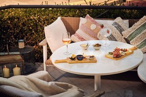 Une table ronde surmontée d’un plateau en marbre blanc, entourée de chaises et de coussins en osier, installée sur une terrasse extérieure avec une haie en arrière-plan.